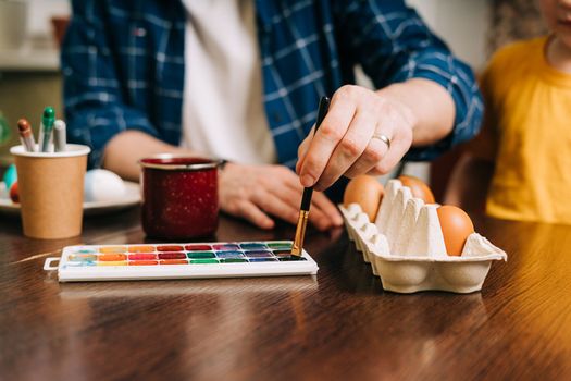 Easter day. Cropped hands of Adult man painting eggs on wooden background. Sitting in a kitchen with crayons. Preparing for Easter, creative homemade decoration.