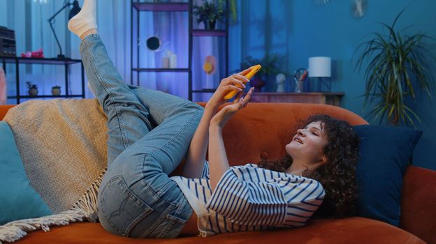 Young woman with curly hairstyle lying on sofa uses mobile phone smiles at night evening home. Girl texting share messages content on smartphone social media applications online watching relax movie