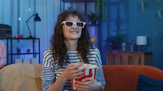 Excited young woman sits on sofa eating popcorn snacks and watching interesting TV serial, sport game, film, online social media movie content at home. Teenager girl enjoying evening entertainment