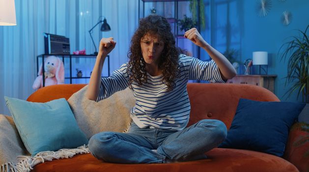 Happy woman with curly lush hairstyle shouting, celebrating success, winning, goal achievement, good news raising fists in gesture I did it, lottery luck at home apartment. Girl in night room on couch
