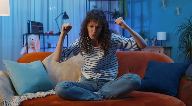 Happy woman with curly lush hairstyle shouting, celebrating success, winning, goal achievement, good news raising fists in gesture I did it, lottery luck at home apartment. Girl in night room on couch