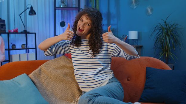 Like. Happy woman with curly hairstyle looking approvingly at camera showing thumbs up, like sign positive something, good great news, positive feedback. Girl sitting on couch at home living room