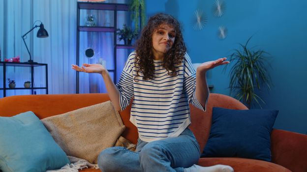 I dont know what to say. Confused young woman feeling embarrassed about ambiguous question, having doubts, no idea, being clueless uncertain at home. Girl at night living room sitting on orange sofa