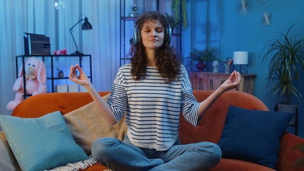Keep calm down, relax. Woman with curly hairstyle at home couch breathes deeply with mudra gesture, eyes closed meditating with concentrated thoughts, peaceful mind, listen music. Girl in evening room