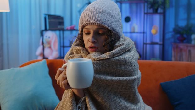 Sick woman wrapped in plaid sit alone shivering from cold on sofa drinking hot tea in unheated apartment without heating due debt. Unhealthy ill girl feeling discomfort try to warm up at evening home