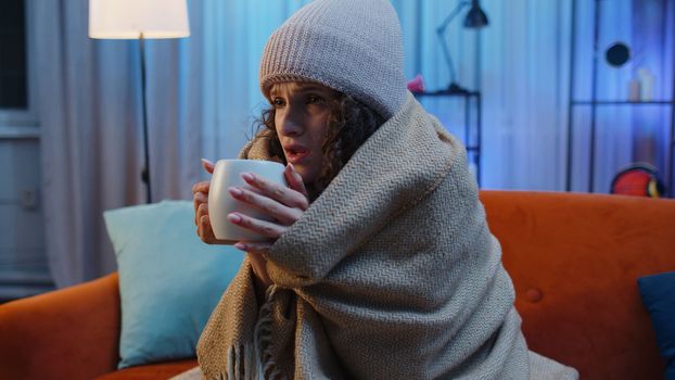 Sick young woman wrapped in plaid sitting, shivering from cold on sofa drinking hot tea in unheated apartment without heating due debt. Unhealthy girl feeling discomfort try to warm up at evening home