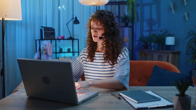 Woman sits at the desk at home wearing headset freelance worker, call center or support service operator helpline, having talk with client or colleague communication support. Girl in evening at home
