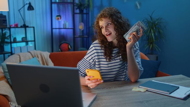 Rich happy young woman counting money cash at smartphone application, making purchasing shopping online, paying for food delivery order sitting at the desk. Girl at modern home office evening room