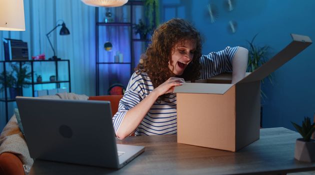 Happy surprised winner woman unpacking delivery parcel at home. Smiling satisfied girl shopper online shop customer opening cardboard box receiving purchase gift headphones by fast postal shipping