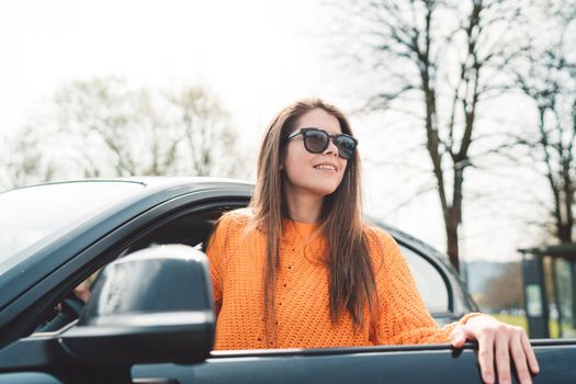 Beautiful young happy smiling caucasian woman driving in her car, wearing sunglasses and an orange sweater. High quality photo