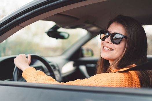 Beautiful young happy smiling caucasian woman driving in her car, wearing sunglasses and an orange sweater. High quality photo
