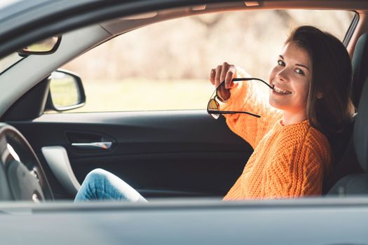 Beautiful young happy smiling caucasian woman driving in her car, wearing sunglasses and an orange sweater. High quality photo