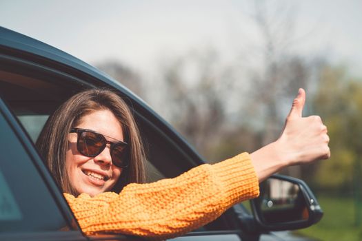 Beautiful young happy smiling caucasian woman driving in her car, wearing sunglasses and an orange sweater. High quality photo