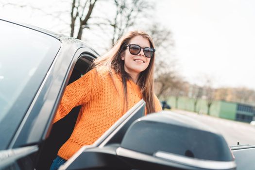 Beautiful young happy smiling caucasian woman driving in her car, wearing sunglasses and an orange sweater. High quality photo