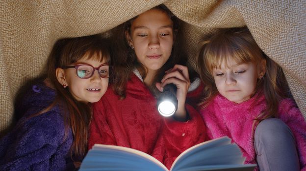 Teen girl and toddler child sisters under blanket plaid covers with flashlight reading interesting fairytale story book using flashlight. Female three siblings children kids friends in wigwam at home