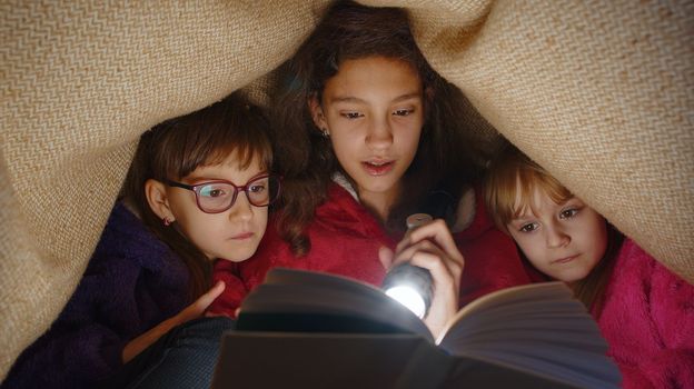 Teen girl and toddler child sisters under blanket plaid covers with flashlight reading interesting fairytale story book using flashlight. Female three siblings children kids friends in wigwam at home