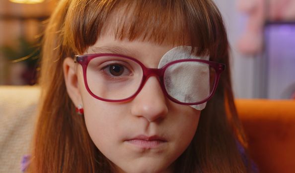 Sick ill toddler girl face looking at camera having protective bandage plaster patch scratch on one eye. Injury trauma blind. Child kid wearing glasses at home in room. Medical eyesight vision therapy