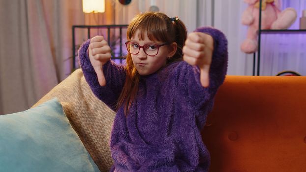Dislike. Upset girl showing thumbs down sign gesture, expressing discontent, disapproval, dissatisfied bad work at modern home apartment indoors. Displeased teen child, kid in living room on sofa