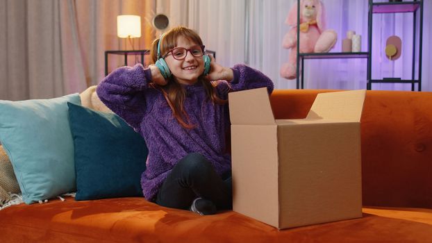 Happy toddler girl unpacking delivery parcel. Smiling satisfied teen child kid shopper, online shop customer opening cardboard box receiving purchase gift by fast postal shipping at home evening room