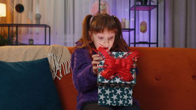 Toddler girl opening gift box with light glow inside, smiling joyfully, looking amazed satisfied with nice present, unexpected birthday surprise. Little child kid at home in evening living play room
