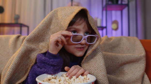 Excited toddler girl in 3D glasses eating popcorn and watching interesting tv serial, sport game, film, online social media movie content. Teen child kid at home in evening living play room on couch