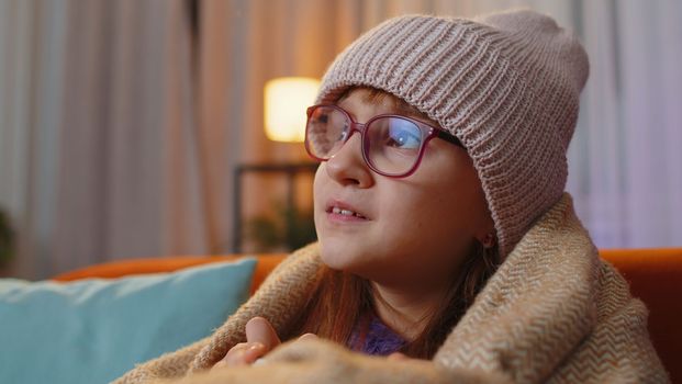 Sick toddler girl wear hat wrapped in plaid sit alone shivering from cold on sofa drinking hot cacao in unheated apartment without heating due debt. Unhealthy kid feeling discomfort try to warming up