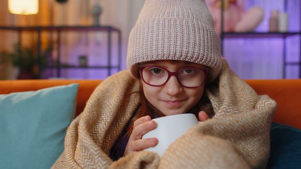 Sick toddler girl wear hat wrapped in plaid sit alone shivering from cold on sofa drinking hot tea in unheated apartment without heating due debt. Unhealthy child feeling discomfort try to warming up
