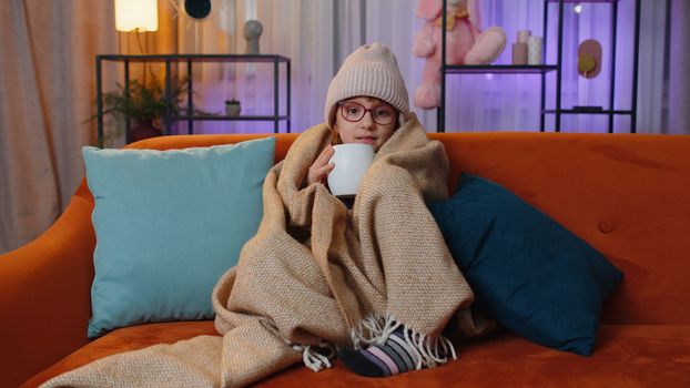 Sick toddler girl wear hat wrapped in plaid sit alone shivering from cold on sofa drinking hot tea in unheated apartment without heating due debt. Unhealthy child feeling discomfort try to warming up