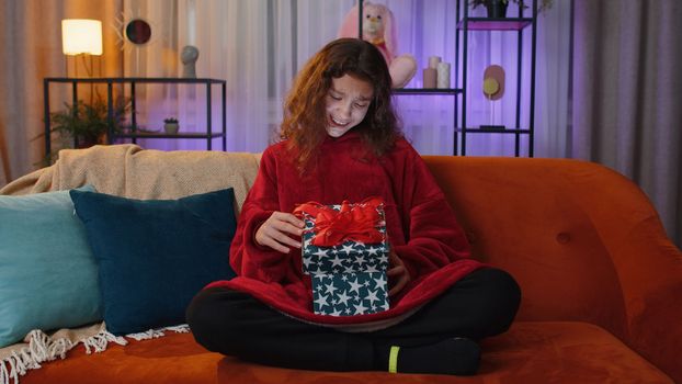 Preteen girl opening gift box with light glow inside, smiling joyfully, looking amazed satisfied with nice present, unexpected birthday surprise. Young child kid at home in evening living play room