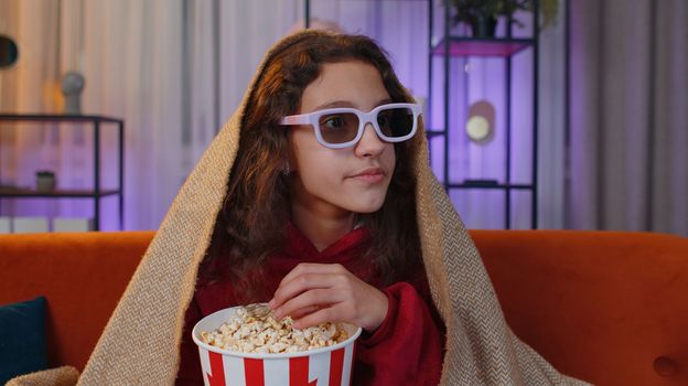 Excited preteen girl in 3D glasses eating popcorn and watching interesting tv serial, sport game, film, online social media movie content. Young child kid at home in evening living play room on couch