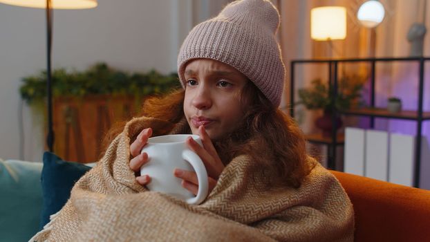 Sick teenager girl wear hat wrapped in plaid sit alone shivering from cold on sofa drinking hot cacao in unheated apartment without heating due debt. Unhealthy kid feeling discomfort try to warming up