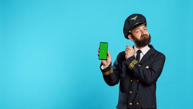 Positive aircrew captain showing phone with greenscreen, holding smartphone with isolated display and chroma key template. Young airplane pilot in uniform looking at mockup copyspace screen.