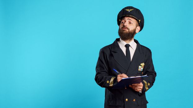 Confident aircrew captain checking list on papers, taking notes with pen and writing flying information. Man working as airplane pilot using clipboard files, wearing aviation uniform and hat.