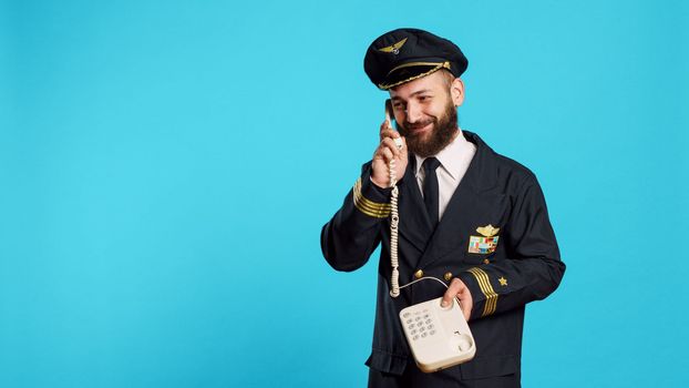 Commercial pilot talking on landline phone call in studio, answering office telephone with cord for remote conversation. Young smiling airliner in uniform chatting on phone line.