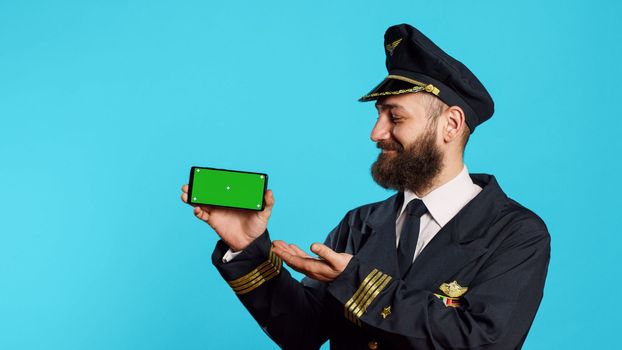 Caucasian man using greenscreen dressed as pilot, showing mobile phone with blank mockup template in studio. Professional flying captain holding phone with isolated chroma key display.