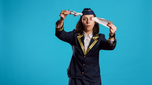 Positive stewardess playing with toy and paper plane, having fun with origami and mini artificial airplane in studio. Happy air hostess wearing flying uniform working in aviation industry.