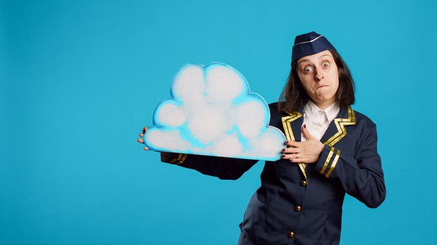 Young stewardess presenting cloud shape billboard, advertising something for aviation industry. Confident air hostess with flying uniform showing isolated cardboard icon, commercial airliner.
