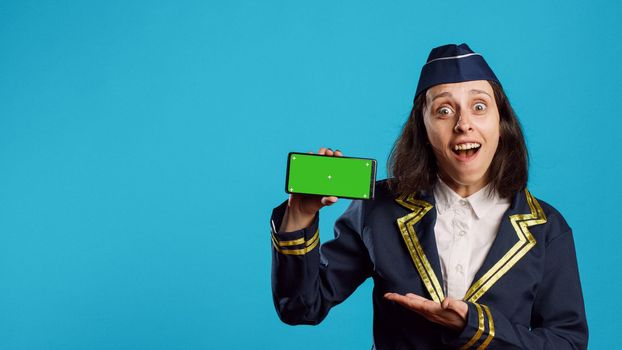 Cheerful air hostess presenting greenscreen on display, working with blank copyspace screen on mobile phone. Smiling airliner using smartphone with isolated chroma key template.