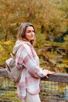 Portrait of cute young woman in casual wear in autumn, standing on bridge against background of an autumn Park and river. Pretty female walking in Park in golden fall. Copy space. smiling girl in the park standing on wooden bridge and looking at the camera in autumn season