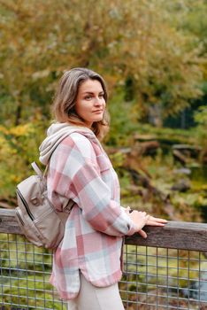 Portrait of cute young woman in casual wear in autumn, standing on bridge against background of an autumn Park and river. Pretty female walking in Park in golden fall. Copy space. smiling girl in the park standing on wooden bridge and looking at the camera in autumn season