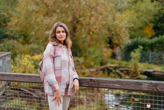 Portrait of cute young woman in casual wear in autumn, standing on bridge against background of an autumn Park and river. Pretty female walking in Park in golden fall. Copy space. smiling girl in the park standing on wooden bridge and looking at the camera in autumn season