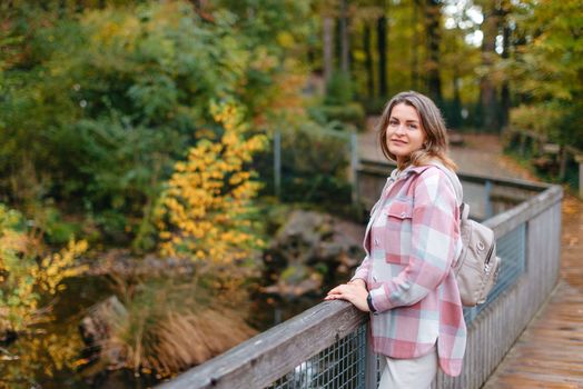 Portrait of cute young woman in casual wear in autumn, standing on bridge against background of an autumn Park and river. Pretty female walking in Park in golden fall. Copy space. smiling girl in the park standing on wooden bridge and looking at the camera in autumn season