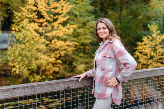 Portrait of cute young woman in casual wear in autumn, standing on bridge against background of an autumn Park and river. Pretty female walking in Park in golden fall. Copy space. smiling girl in the park standing on wooden bridge and looking at the camera in autumn season