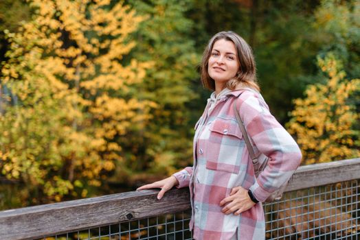 Portrait of cute young woman in casual wear in autumn, standing on bridge against background of an autumn Park and river. Pretty female walking in Park in golden fall. Copy space. smiling girl in the park standing on wooden bridge and looking at the camera in autumn season