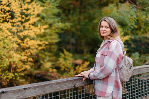 Portrait of cute young woman in casual wear in autumn, standing on bridge against background of an autumn Park and river. Pretty female walking in Park in golden fall. Copy space. smiling girl in the park standing on wooden bridge and looking at the camera in autumn season