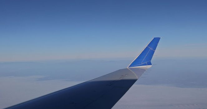An airplane wing flying above clouds as seen through window against background of fluffy clouds