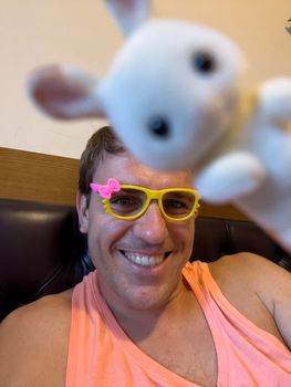 Man in toy glasses sits on a bed with a toy hare. High quality photo