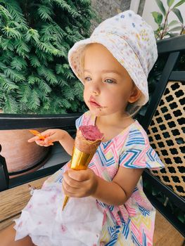 Little girl is sitting on a bench near a tree with popsicles in a cone. High quality photo