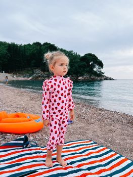 Little girl stands on a blanket on a pebble beach and looks into the distance. High quality photo