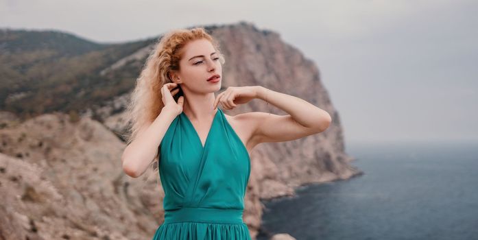 Side view a Young beautiful sensual woman in a mint long dress posing on a volcanic rock high above the sea during sunset. Girl on the nature on overcast sky background. Fashion photo